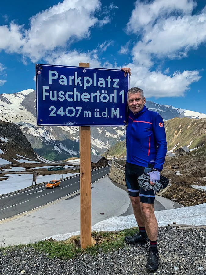 Scenic cycling journey from Salzburg to Grado: a person stands next to a signpost reading &apos;Parkplatz Fuschertörl 1 2407 m&apos;, with snow-capped mountains in the background